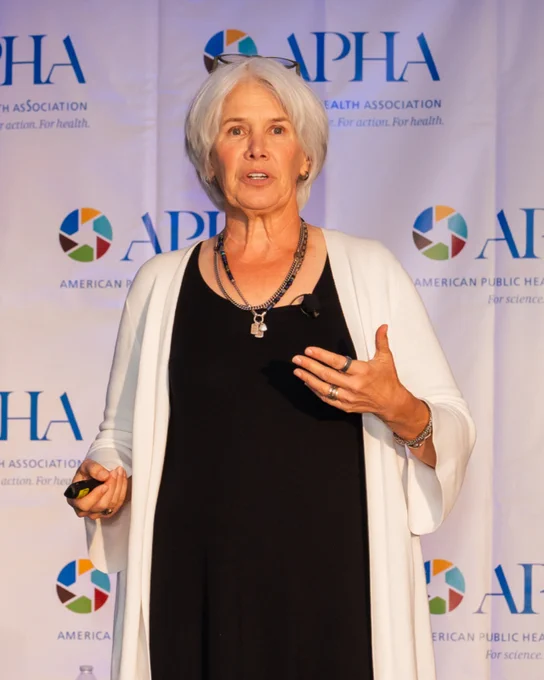 A confident speaker presents at a public health conference. The banner in the background displays the American Public Health Association (APHA) logo.