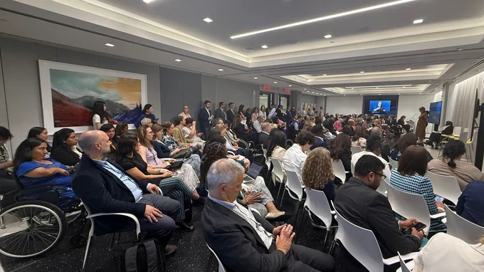 A large, bright conference room filled with many seated attendees facing the front. A speaker stands near a screen, addressing the group, while a sign language interpreter appears on a display. Some people take notes or use phones, and a few use wheelchairs. The room is full.