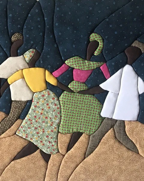 Textile artwork of four stylized figures dancing hand in hand beneath a crescent moon and star-filled night sky. The figures wear patterned dresses and headwraps in floral, green, pink, yellow, and white fabrics, moving across rolling sandy hills made from textured cloth.
