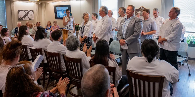 A group of chefs and a person in a suit stand in front of an audience seated in a well-lit room, with some attendees applauding.
