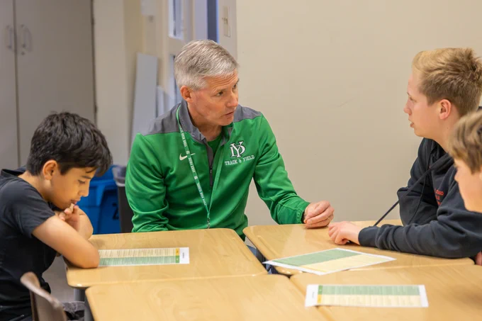 A teacher in a green jacket engages with three students seated at desks in a classroom. The mood is focused and collaborative.