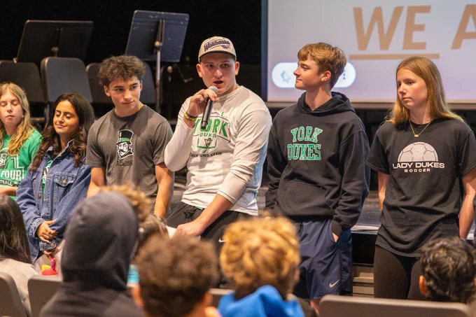 Students gathered on stage for a school event. One speaks into a microphone while others stand nearby. The mood is engaging and lively.