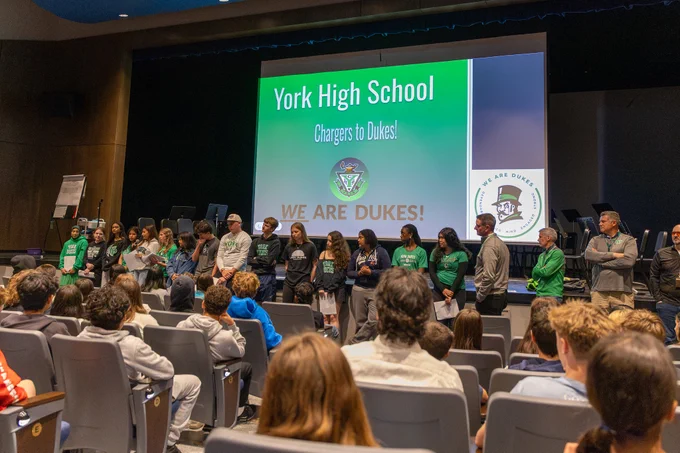 Students and staff stand on stage for a presentation at York High School, celebrating school spirit with a large screen displaying the message 'We Are Dukes'. The atmosphere is engaging and inclusive.