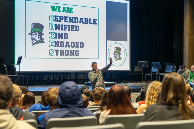 A speaker presents to students on the values of being Dependable, Unified, Kind, Engaged, and Strong, symbolized by the acronym 'DUKES'. The event takes place in an auditorium setting with a focus on community and school spirit.
