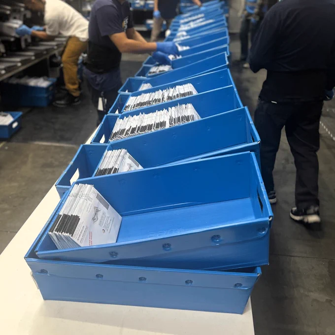 A row of bins in a vote counting center. The bins are partially filled with ballots. There are people working in the background.