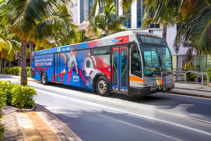 City bus with colorful soccer-themed wrap featuring a player kicking a ball, driving down a palm-lined street in a sunny urban area.