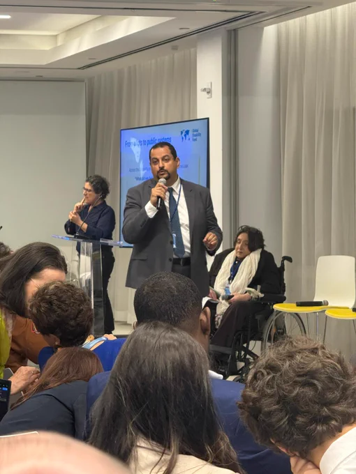 Ahmed Ghanem stands at the front of a conference room, holding a microphone and addressing a seated audience. A sign language interpreter stands nearby at a clear podium. Behind them, a large screen shows slides. To the side, a panel includes Dr. Ola Abualghaib  seated in a wheelchair. Rows of attendees face forward.