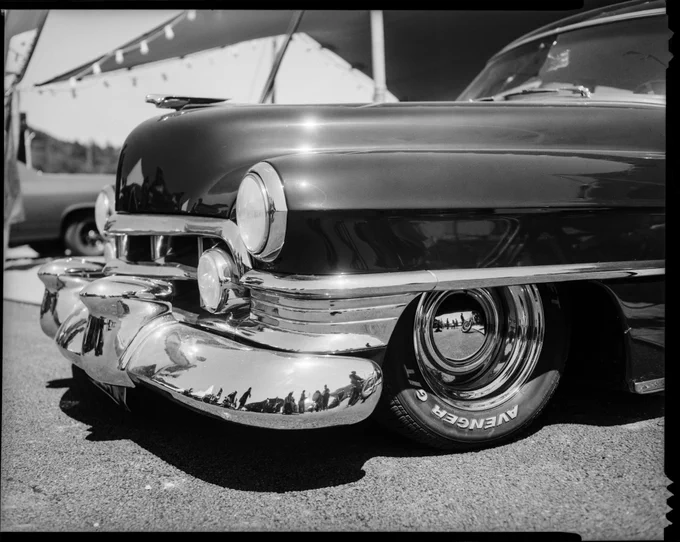 a black and white large format photograph of an early 1950s Chevrolet Deluxe Coupe, showing the front of the car with a lot of chromed part under the headlight, the hood and front wheel and a bit of the windshield. The image is shot so our eyes are at hood level.