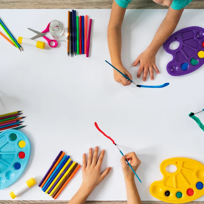 Two children drawing on a large sheet of paper with crayons, markers, and paint palettes laid out around them.