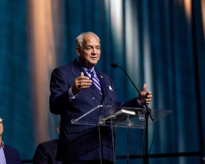 A man in a suit speaks at a transparent podium against a blue curtain backdrop, conveying a formal and professional setting.