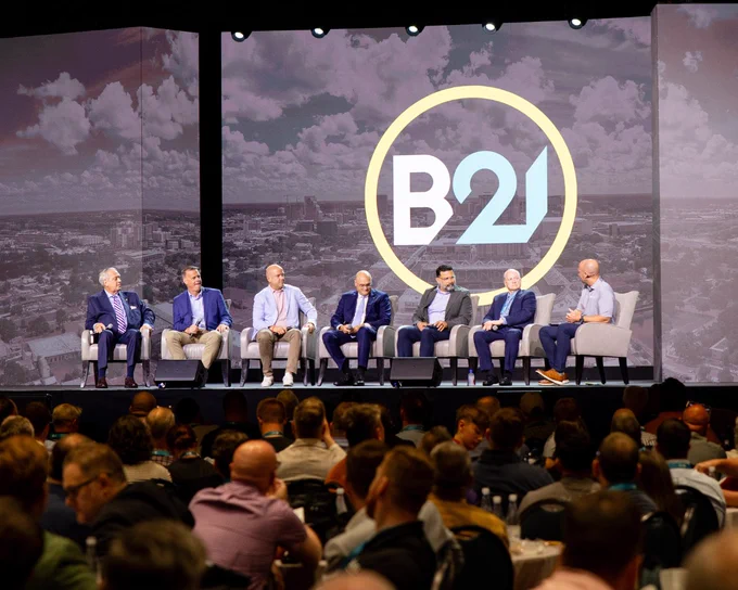A group of seven men in formal attire are seated on stage for a panel discussion at the B21 Conference. The background displays a cityscape with a large B21 logo.