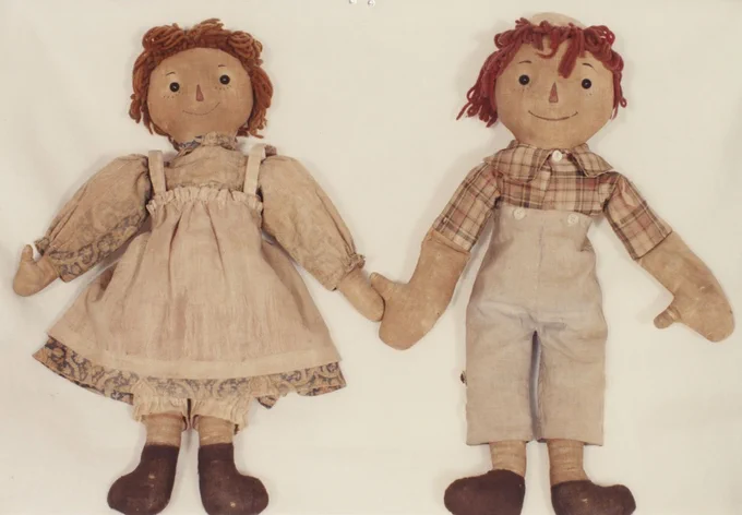 The original Raggedy Ann and Andy cloth dolls stand side-by-side, holding hands. Ann (left) has red yarn hair, wearing a dress over patterned long sleeves. Andy (right) has red yarn hair under a cap, wearing a plaid shirt and light beige overalls. Both have stitched faces and dark brown feet.