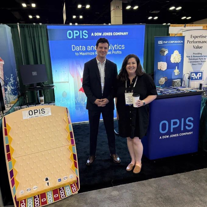 A person stands next to another at an OPIS and Dow Jones Company booth. There's a vertical board with slots labeled OPIS in front, and banners with text about data analytics. The booth has displays and promotional materials.
