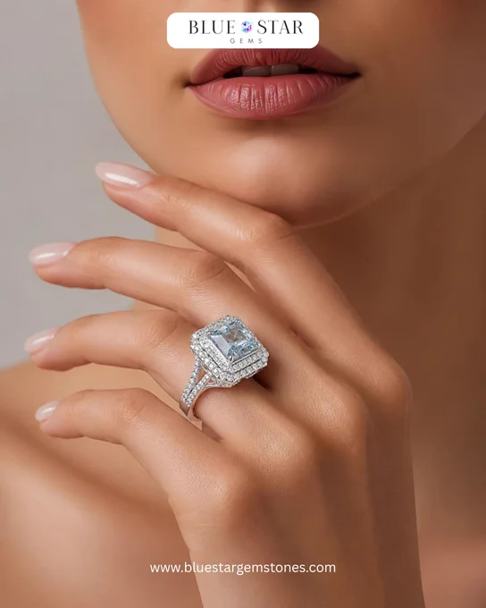 A close-up portrait of a woman wearing a square aqua-blue gemstone ring surrounded by a diamond halo setting. The ring is set on a silver band, and the model’s hand is gently placed near her face, highlighting the ring’s brilliance and elegant design against a soft neutral background.