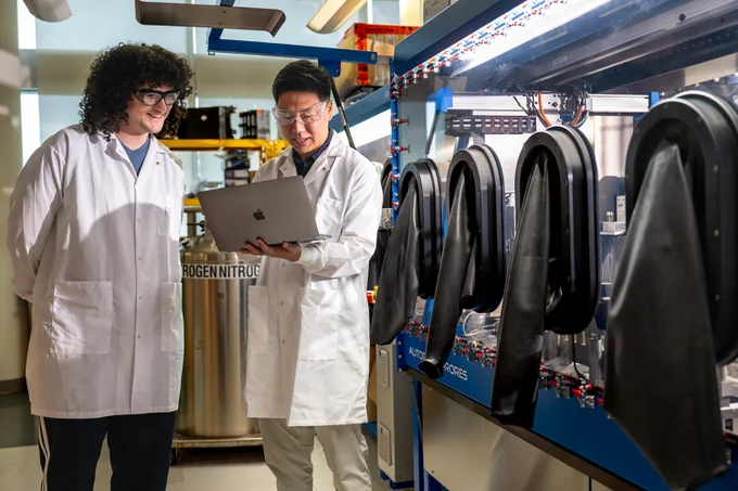 Two scientists in white lab coats discussing data on a laptop inside a laboratory.