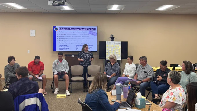Educators in a circle and at tables listen to Kris Kampovitz, standing in the center, talk about the slide show behind her. The slide is titled "Collaborative Team Roles - Who Will You Be?"