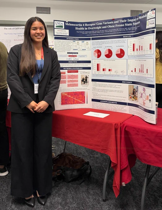 A person stands smiling in front of a presentation board titled "Melanocortin 4 Receptor Gene Variants and Their Impact on Metabolic Health in Overweight and Obese Fresno State Students." The board, featuring the USDA logo, displays charts, graphs, and text. They are dressed in business attire, and there is a bag on the floor beside them.