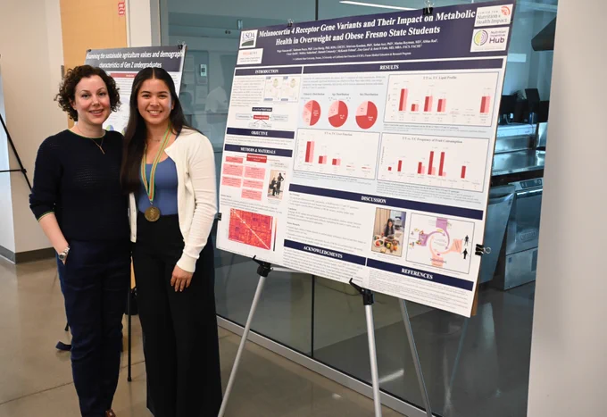 Two individuals stand beside a scientific poster presentation in an indoor setting. The poster is titled "Melanocortin 4 Receptor Gene Variants and Their Impact on Metabolic Health in Overweight and Obese Fresno State Students" and includes graphs and text. One person wears a medallion.
