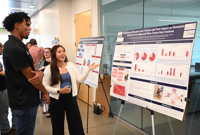 Two individuals are discussing research posters displayed on stands in a well-lit room. One poster is titled "Maternal and Paternal Care Values and Their Impact on Metabolic Health in Overweight and Obese Fresno State Students," featuring various charts and graphs. Other people are observing additional posters in the background.