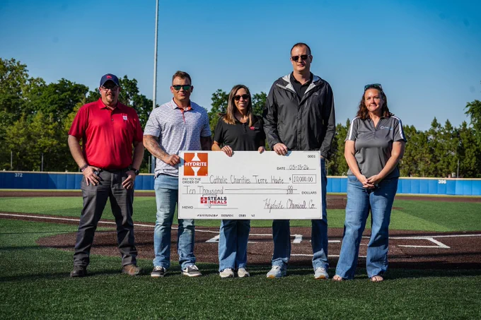 Five people standing on a baseball field holding a large check made out to Catholic Charities Texas for ten thousand dollars.