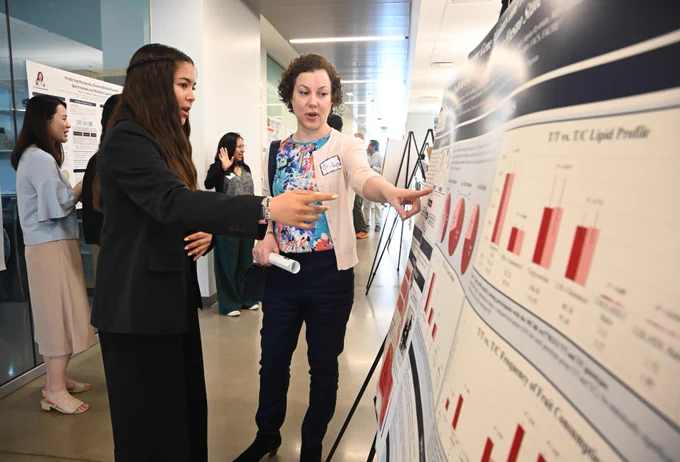Two people are engaged in discussion in front of a large scientific poster displaying bar graphs and charts. The setting appears to be a conference or academic presentation. Other individuals are interacting in the background.
