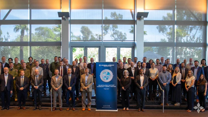 Large group of professionals posing indoors behind a blue banner with organizational banner and text about the General Atomics Aeronautical Systems, Inc. second MQ-9B Global Support Community Meeting.
