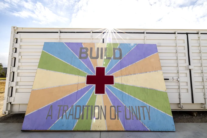 A colorful mural leaning against a white shipping container features a red cross at the center, surrounded by radiating stripes in various pastel colors. The words "BUILD" and "A TRADITION OF UNITY" are prominently displayed. Sunlight beams over the top edge, creating a bright and hopeful atmosphere.