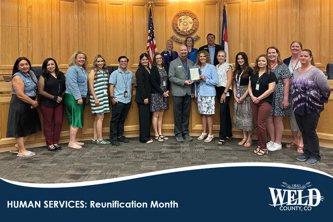 Group of Weld County Human Services staff posing in a wood-paneled room holding a certificate for Reunification Month.