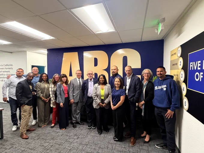 Group of diverse professionals posing in front of a large blue wall with bold gold letters 'ABC' in an office setting.