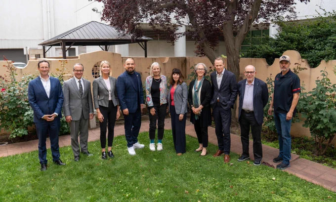 Group of ten professionally dressed people standing outdoors on grass with trees and a building in the background.