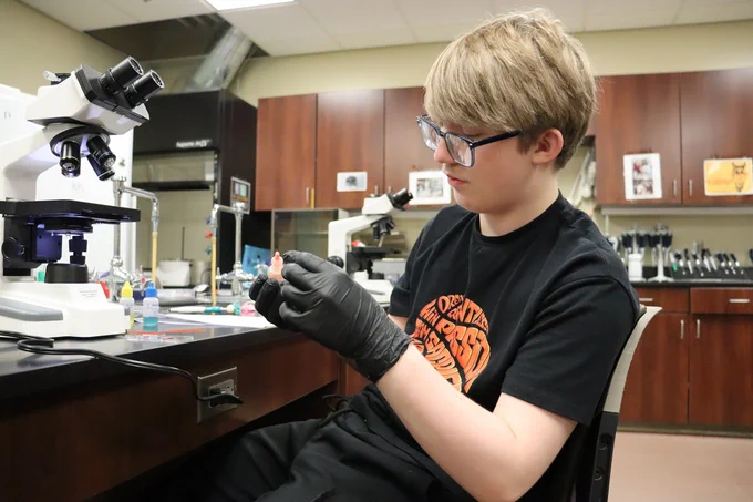 Student wearing black gloves carefully handling a small object in a science lab filled with microscopes and lab equipment.