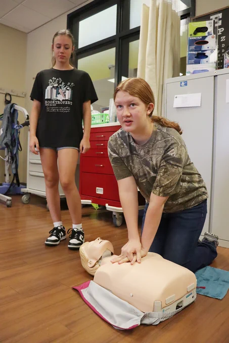 Person practicing CPR chest compressions on a training mannequin in a clinical setting while another watches.
