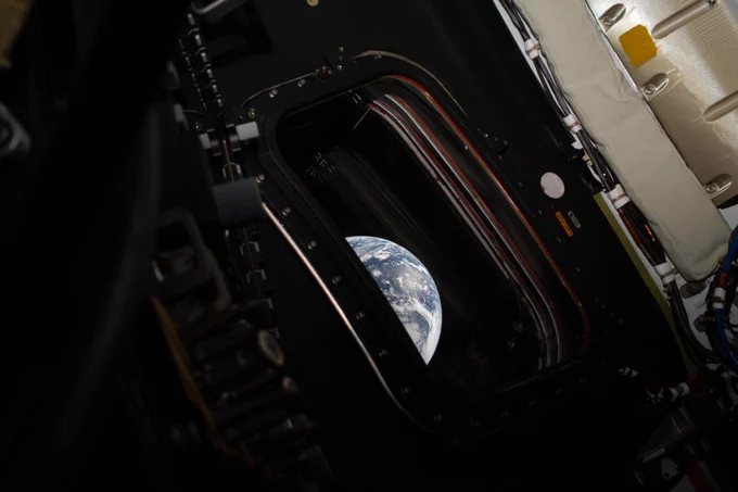 A view of Earth through a spacecraft window, showing the blue-and-white planet partially framed by the dark interior structure, cables, and hardware inside the spacecraft.
