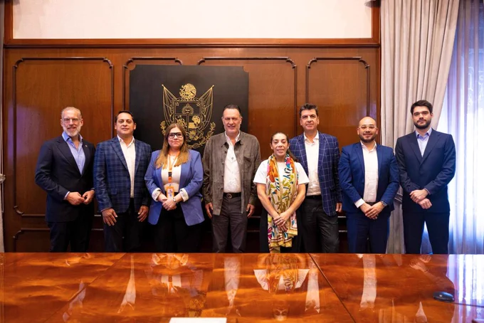 Ocho personas vestidas formalmente posan frente a una mesa de madera brillante en una sala con paredes de madera y cortinas. Al fondo, hay un escudo en la pared.