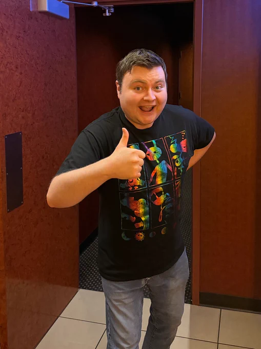 Krazy Ken standing outside the door to a movie theater auditorium. He's smiling and giving a thumbs up while wearing a black t-shirt with rainbow-colored characters from The Amazing Digital Circus cast.