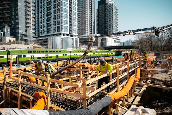 Des ouvriers travaillent sur un chantier de construction, avec de hauts immeubles en arrière-plan. Un train GO Transit vert passe au second plan. On aperçoit des matériaux et de l'équipement de construction autour du site.