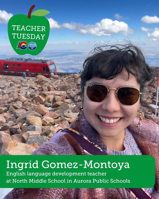 A woman stands on a mountain with the Pikes Peak train behind her and a rocky ground landscape. She wears a dark pink jacket and round sunglasses. Text overlay reads: Teacher Tuesday - Ingrid Gomez-Montoya, English language development teacher at North Middle School in Aurora Public Schools.