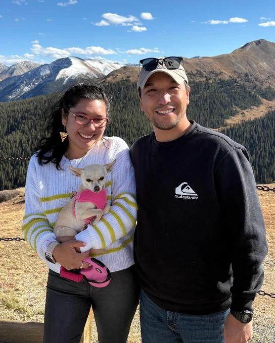 Two people stand outdoors smiling, with mountains in the background under a blue sky. One person is holding a small white dog wearing a pink outfit. They are both casually dressed, and both are wearing sunglasses.