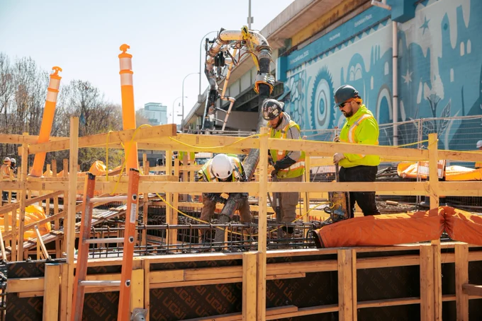 Travailleurs sur un chantier de construction avec de grands bâtiments dans l’arrière-plan. Un train vert de GO Transit passe à une distance médiane. Des matériaux de construction et de l’équipement sont visibles autour du site.