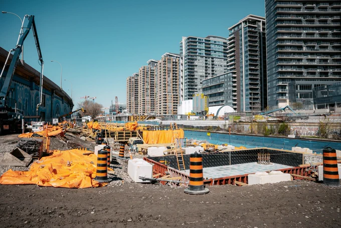 Un chantier de construction avec des travailleurs et des machines près d’un immeuble de grande hauteur. Il y a des cadres en bois avec plusieurs travailleurs en équipement de sécurité orange. Les machines avec un long bras s’étendent sur le site. À l’arrière-plan, une murale est visible sur un mur, et une partie de la Tour CN peut être vue.