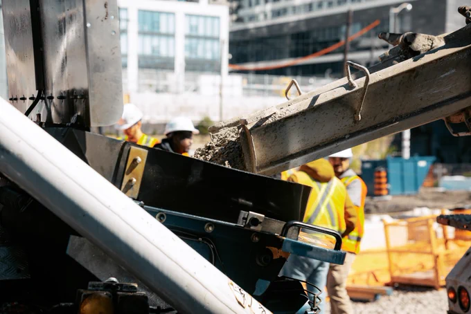 Un groupe de travailleurs sur un chantier de construction construit un coffrage en bois. Ils portent de l’équipement de sécurité, y compris des casques et des gilets réfléchissants. À l’arrière-plan, une grande murale est peinte sur un mur sous un passage supérieur. La scène est occupée par des activités de construction.