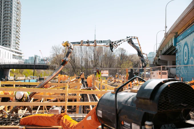 Un groupe de travailleurs sur un chantier de construction construit un coffrage en bois. Ils portent de l’équipement de sécurité, y compris des casques et des gilets réfléchissants. À l’arrière-plan, une grande murale est peinte sur un mur sous un passage supérieur. La scène est occupée par des activités de construction.