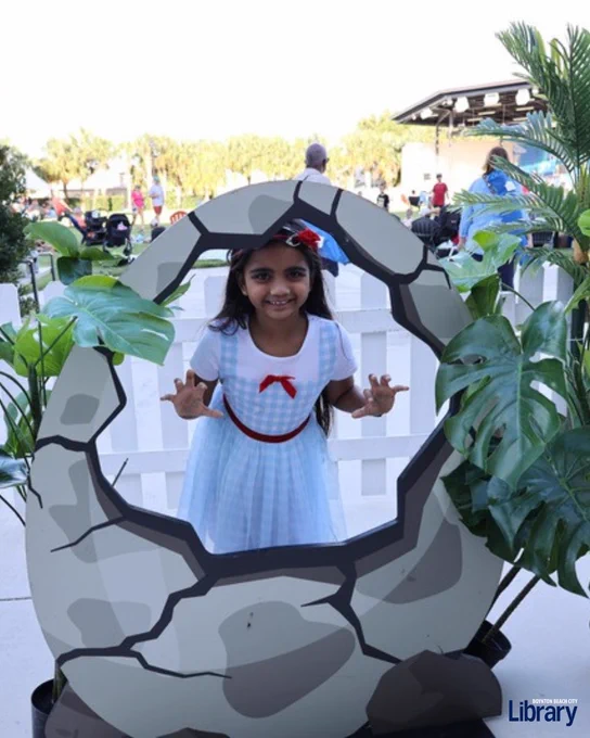 Young girl in a blue dress with a red bow posing behind a large cracked stone egg prop outdoors.