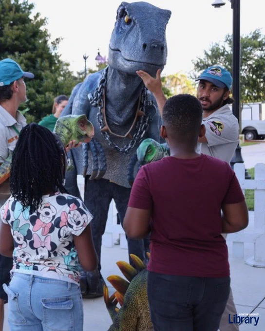 Children interact with a person in a realistic blue dinosaur costume outdoors near a white fence and trees.