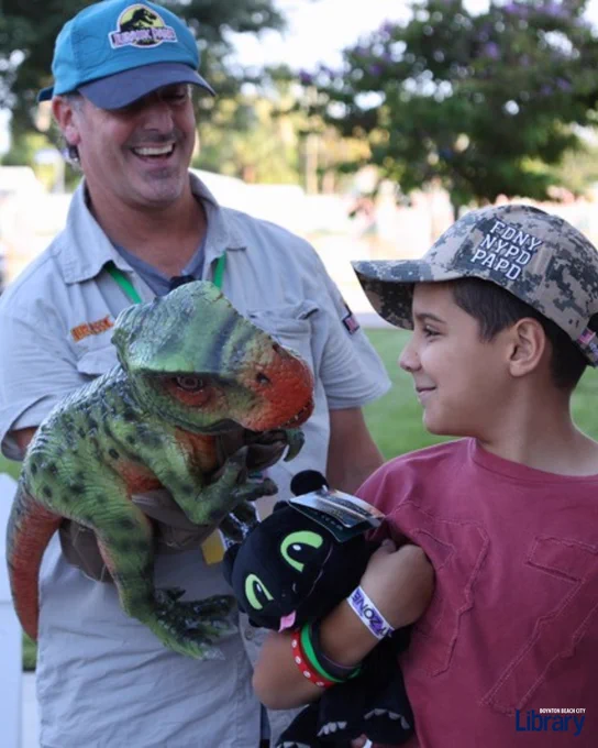 Child wearing a camouflage cap holding a black plush toy while looking at a person holding a realistic green and orange dinosaur puppet outdoors.