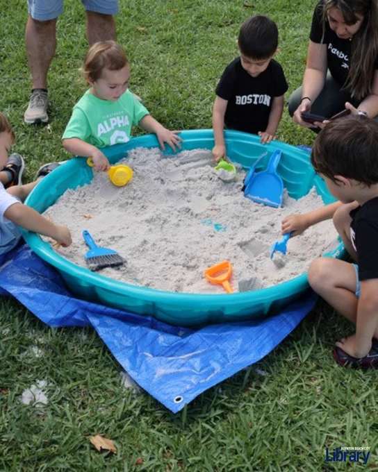 Children playing with colorful shovels and rakes in a blue plastic sandbox filled with sand on a grassy lawn.