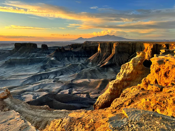 Sunset casting golden light over rugged canyon cliffs and layered rock formations under a partly cloudy sky.