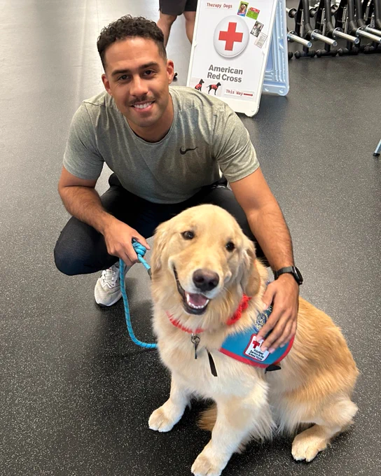 A person is smiling and crouching next to a Golden Retriever wearing a therapy dog vest, featuring an American Red Cross logo. There is a sign in the background displaying the American Red Cross logo and text.