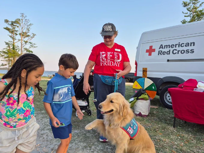 A group of individuals is outdoors near an American Red Cross vehicle. One person wearing a "RED Friday" shirt stands by a golden retriever with a service vest. Two children are interacting with the dog, which is lifting a paw. A colorful spinner and some tables are visible in the background.