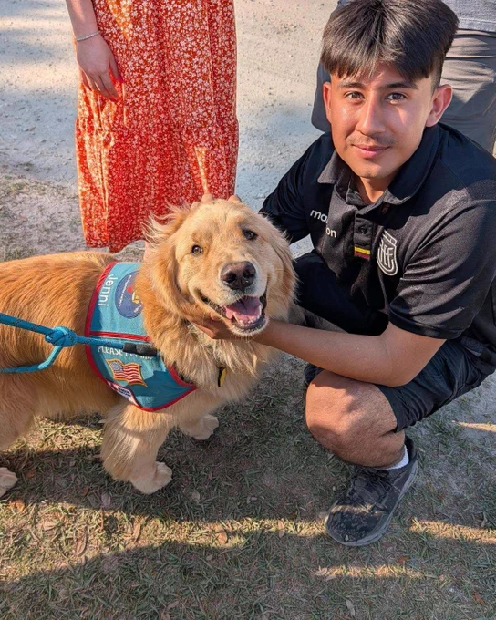 There is a person crouching next to a golden retriever wearing a blue service dog vest with an American flag patch. The person is holding the dog's face gently and smiling. There is another person partially visible, wearing a red and white patterned skirt. The setting appears to be outdoors on a sunny day.